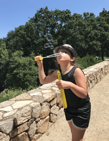 A young girl stands on a sunny stone pathway, blowing colorful bubbles into the air while holding a bottle of bubble solution. Surrounded by lush green trees under a bright blue sky, she enjoys a playful summer moment outdoors.